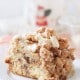 A close-up of a slice of crumbly coffee cake on a white plate showcases the perfect Coffee Crumb Cake recipe. The cake boasts a dense texture with crumbled topping and drizzled icing, while a blurred mug with a festive design adds charm in the background.