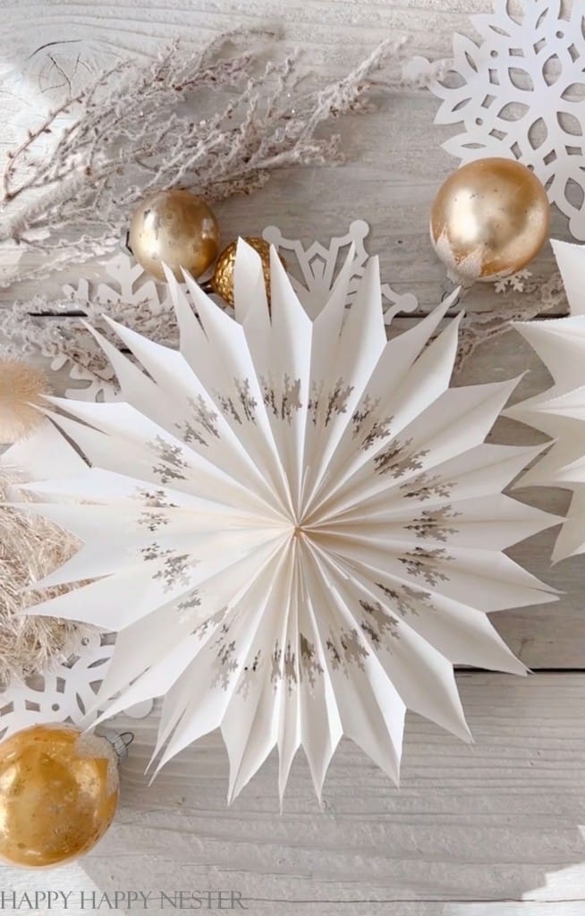 A handmade paper snowflake decoration is surrounded by gold baubles, dried twigs, and additional paper snowflakes on a wooden surface. This charming arrangement of Christmas crafts for adults creates a wintery, festive atmosphere.