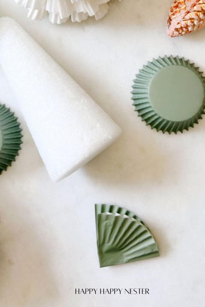 Craft materials are arranged on a surface, including green cupcake liners in various states of folding, a white foam cone, and a pine cone. The text "HAPPY HAPPY NESTER" is at the bottom.