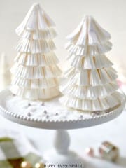 Two decorative Christmas trees crafted with a Paper Christmas Tree DIY technique using stacked white paper cupcake liners are displayed on a white cake stand. The background features a soft focus with subtle festive elements.