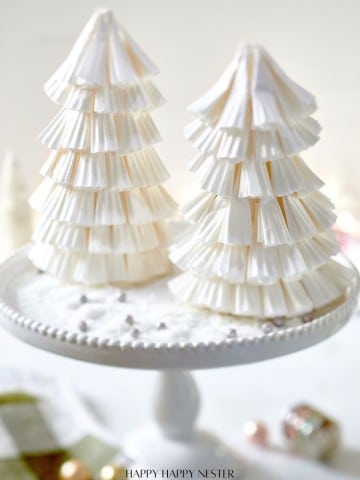 Two decorative Christmas trees crafted with a Paper Christmas Tree DIY technique using stacked white paper cupcake liners are displayed on a white cake stand. The background features a soft focus with subtle festive elements.