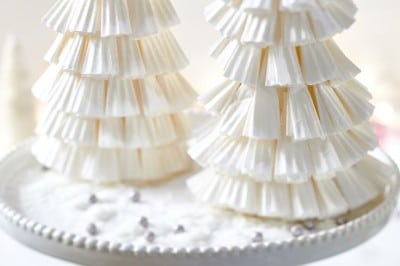 Two decorative Christmas trees crafted with a Paper Christmas Tree DIY technique using stacked white paper cupcake liners are displayed on a white cake stand. The background features a soft focus with subtle festive elements.
