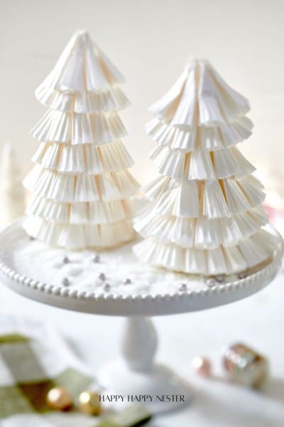 Two decorative Christmas trees crafted with a Paper Christmas Tree DIY technique using stacked white paper cupcake liners are displayed on a white cake stand. The background features a soft focus with subtle festive elements.