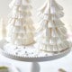 Two decorative Christmas trees crafted with a Paper Christmas Tree DIY technique using stacked white paper cupcake liners are displayed on a white cake stand. The background features a soft focus with subtle festive elements.