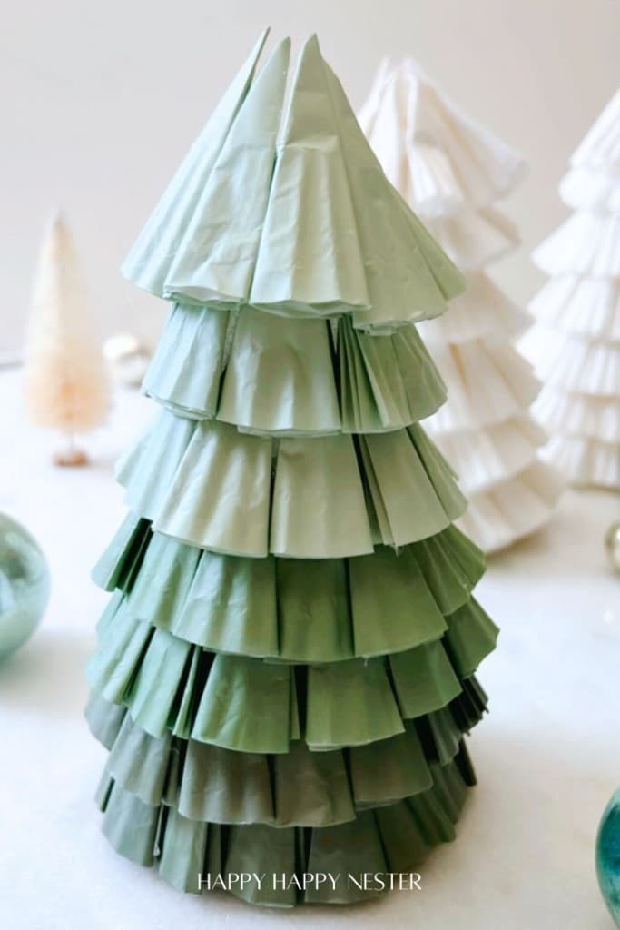 A green paper Christmas tree decoration, reminiscent of a DIY project, features layered, fan-shaped sections. It stands on a white surface with blurred white and cream trees in the background, exuding a festive holiday vibe.