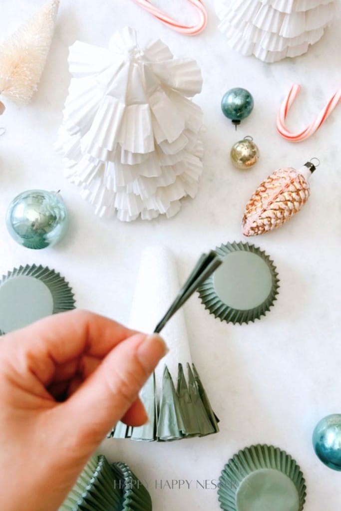 A hand holding a piece of paper next to green cupcake liners, with white paper trees, Christmas ornaments, candy canes, and pine cone ornaments scattered on a light background.