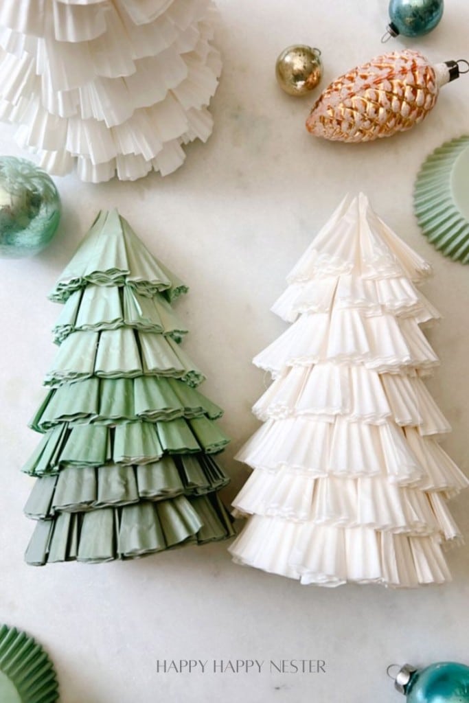 Two handmade Christmas trees crafted from paper cupcake liners, one green and one white, are displayed on a light surface. They are surrounded by decorative ornaments, such as round teal and gold baubles, and a pinecone-shaped ornament.