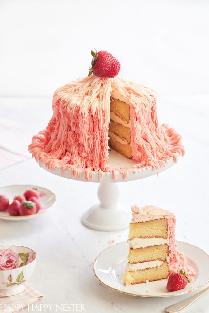 A layered cake with pink whipped frosting sits on a white pedestal, serving as delightful cake inspiration. A whole strawberry crowns the creation, while a slice is elegantly placed on a nearby plate. In the background, a small bowl of strawberries and a floral teacup complete the charming scene.