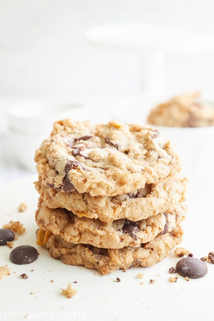 A stack of four chocolate chip cookies, touted as the best Christmas cookies, sits on a white surface. Crumbs and a few chocolate chips are scattered around. The background is softly blurred, highlighting these festive treats in the foreground.