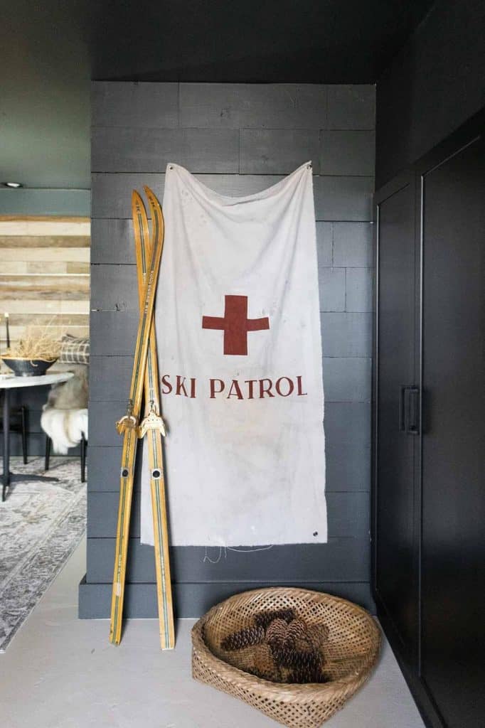 A pair of vintage yellow wooden skis are leaned against a wall alongside a white "Ski Patrol" banner with a red cross. Below, air dry clay pinecones fill a basket. The setting, reminiscent of a cozy clay house, blends modern and rustic design elements indoors.