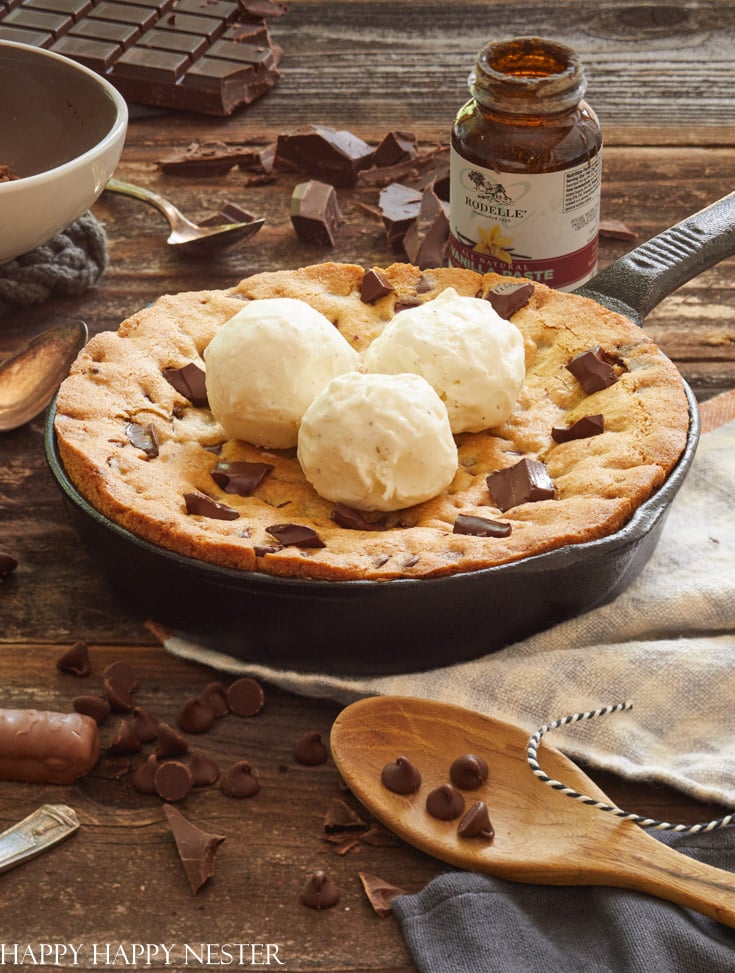 A skillet cookie, a festive chocolate Christmas treat, sits on a wooden table topped with three scoops of vanilla ice cream. The decadent dessert is sprinkled with chocolate chunks, surrounded by chocolate chips, a wooden spoon, a jar of hazelnut spread, and pieces of chocolate bars.