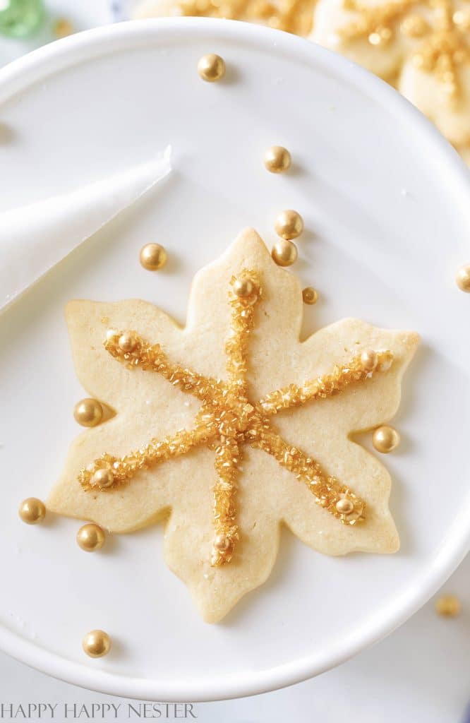 A maple leaf-shaped cookie, one of the best Christmas cookies, is elegantly decorated with gold sugar and sprinkles on a white plate. Additional gold sprinkles are scattered around, and a frosting bag rests in the top left corner.