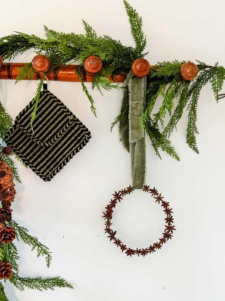 A festive wall decoration featuring green pine branches, a striped black and gold pouch, and a hanging star anise wreath adorn a wooden rack with pegs. Nestled among the branches is a charming air dry clay house, adding to the natural pinecone-studded holiday display.