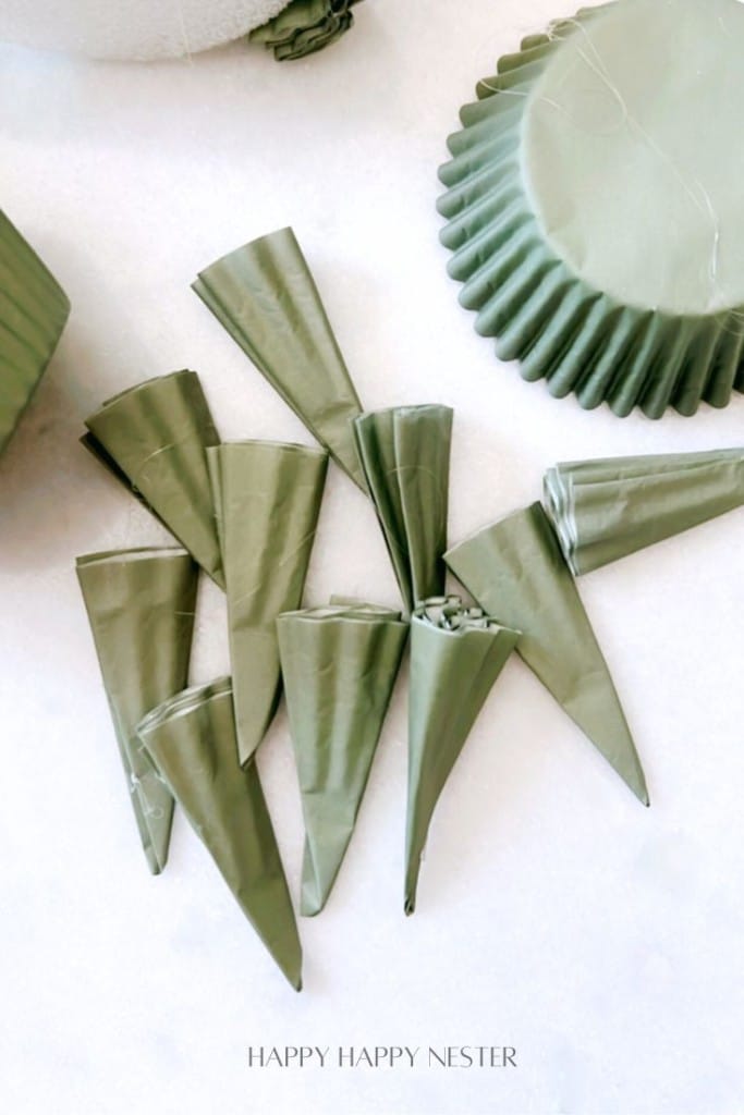 A collection of olive green, folded paper cones resembling a DIY Christmas tree are neatly arranged on a light background. Nearby, a stack of matching pleated paper forms is partially visible.