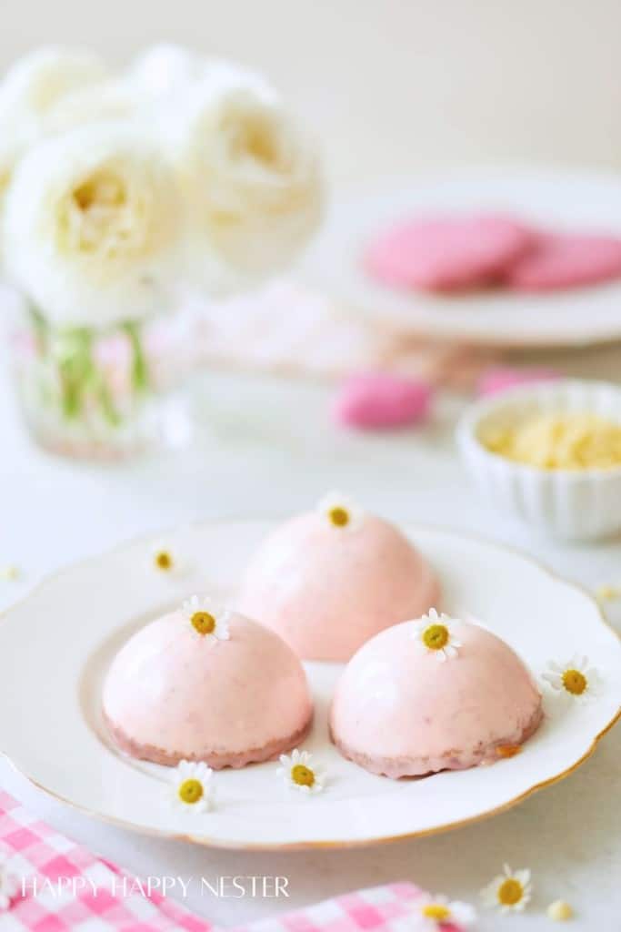 A plate of three round pink desserts, offering cake inspiration, is topped with small white flowers on a table. In the background, there's a jar of white roses, a bowl of cream, and some pink cookies, creating a soft, elegant setting.