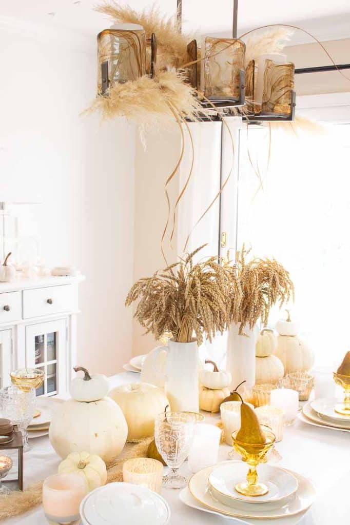 A dining table adorned with fall table decor (Amazon) features white pumpkins, wheat stalks in vases, and golden accents. A modern chandelier with pampas grass floats above. Elegant glassware and candles complete the setting, creating a warm, rustic atmosphere.