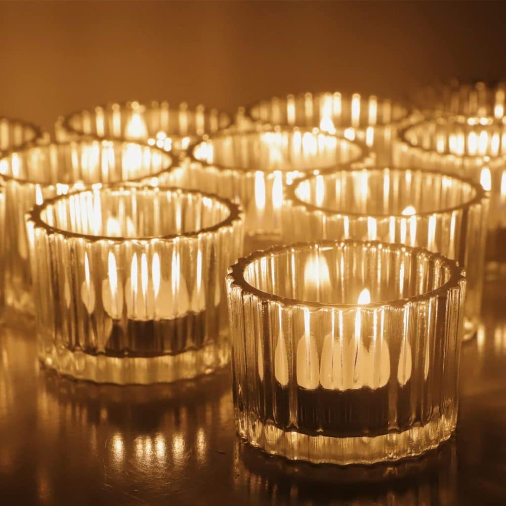 A warm, golden glow emitted by numerous lit candles placed in ribbed glass holders on a reflective surface. Among them sits an air dry clay house, adding charm as the ambient lighting creates a cozy and inviting atmosphere.