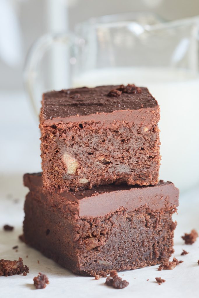 Two square pieces of chocolate brownie stacked on a white surface, with a blurred glass pitcher of milk in the background. These rich Christmas treats are dense with a smooth top layer. Chocolate crumbs are artfully scattered around them, adding to their festive allure.