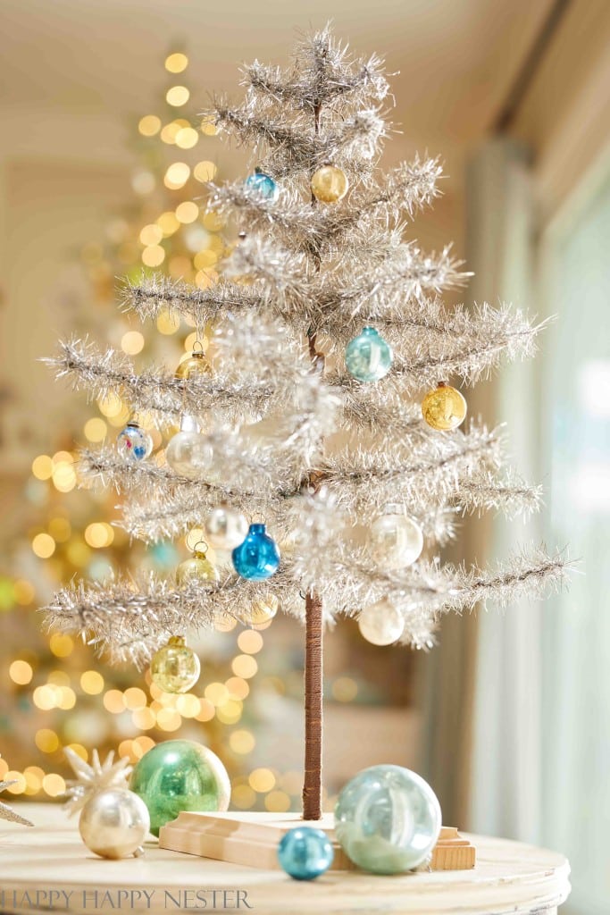 A small silver tinsel Christmas tree, perfect for Christmas crafts for adults, is adorned with colorful ornaments like blue, gold, and white baubles. In the background, a larger tree with golden lights is softly blurred. Decorated baubles are artfully placed at the base of the small tree.