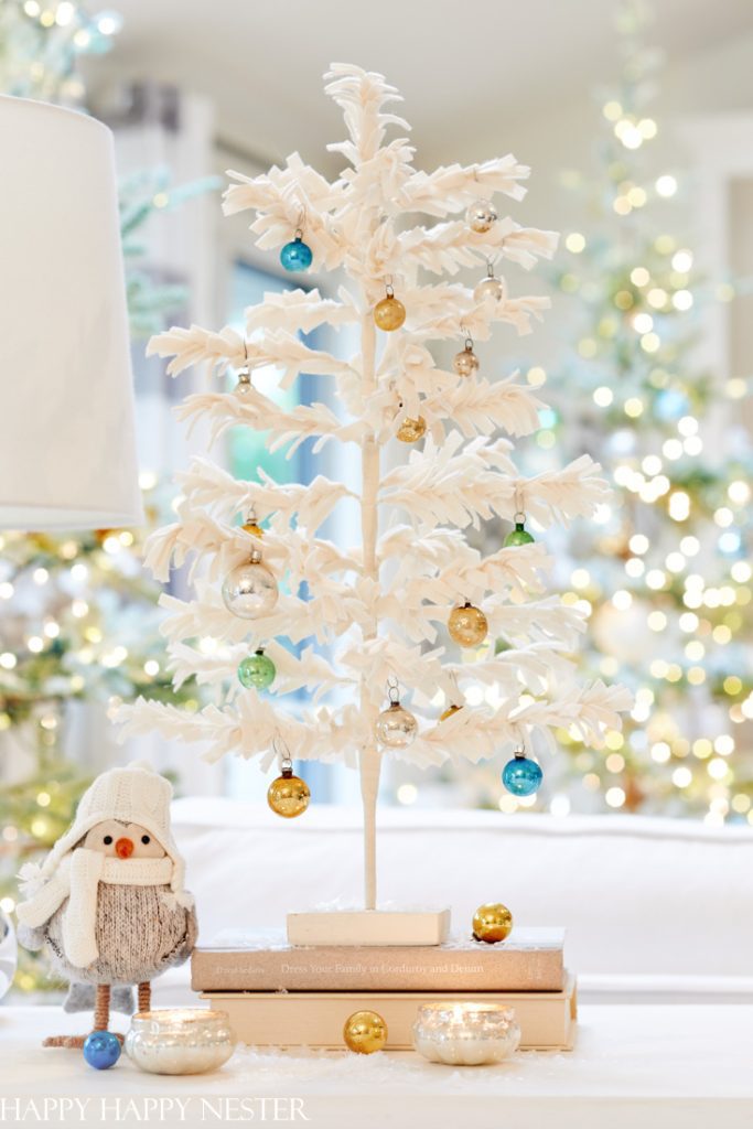 A small, white, feathery Christmas tree adorned with blue, silver, and gold baubles sits atop a stack of books—a delightful example of Christmas crafts for adults. A cute, plush penguin toy wearing a knitted hat and scarf is placed nearby. Warm lights twinkle softly in the blurred background.