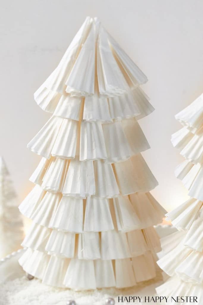 A festive paper Christmas tree DIY, crafted from stacked white cupcake liners, exhibits a tiered, layered appearance. Set against a neutral background, this decorative piece adds a unique charm to any holiday setting.