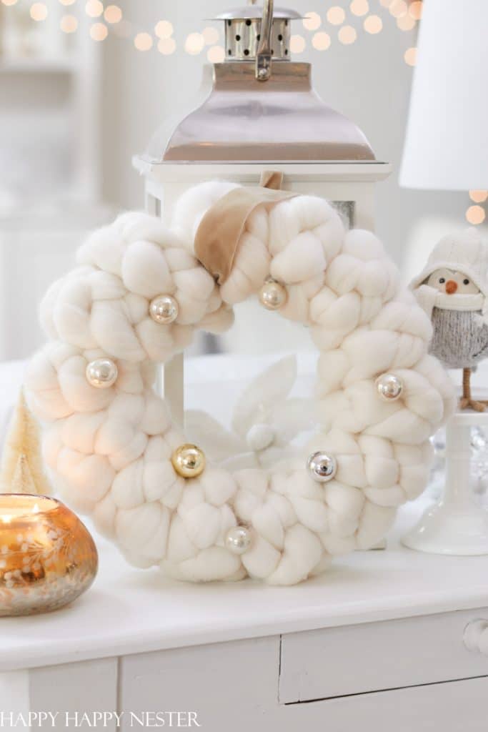 A cozy white wreath made of chunky knit wool, adorned with shiny gold and silver baubles, hangs on a white background. Nearby, a decorative lantern, small ornaments, and a figure wearing a winter hat enhance the festive atmosphere. It's an ideal project for those who enjoy Christmas crafts for adults.