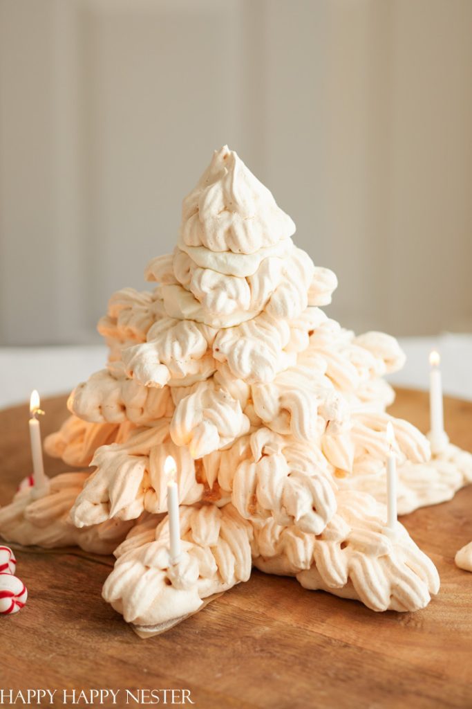 A Christmas tree-shaped meringue dessert on a wooden board, resembling a snowy tree and perfect for holiday gatherings. Four small candles glow at the base. The light and fluffy meringue, with its creamy color, offers a delightful addition to your favorite Christmas treat recipes.