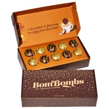 Open and closed boxes of "BoniBombs" hot chocolate bombs are displayed. The box design is dark brown with orange accents and gold foil, showcasing text about enjoying hot chocolate. The bombs are covered in gold foil with chocolate drizzle.