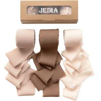 A set of three rolls of fabric ribbon in shades of brown and beige, placed next to a cardboard box with a transparent window displaying the brand name "JEDIA." The ribbons have frayed edges and are artistically arranged.