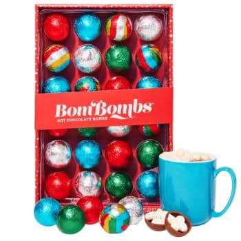 A box labeled "BomBombs Hot Chocolate Bombs" contains colorful, foil-wrapped chocolate spheres. Beside the box, a blue mug is filled with marshmallow-topped hot chocolate, with two open chocolate bombs revealing cocoa and marshmallow filling.