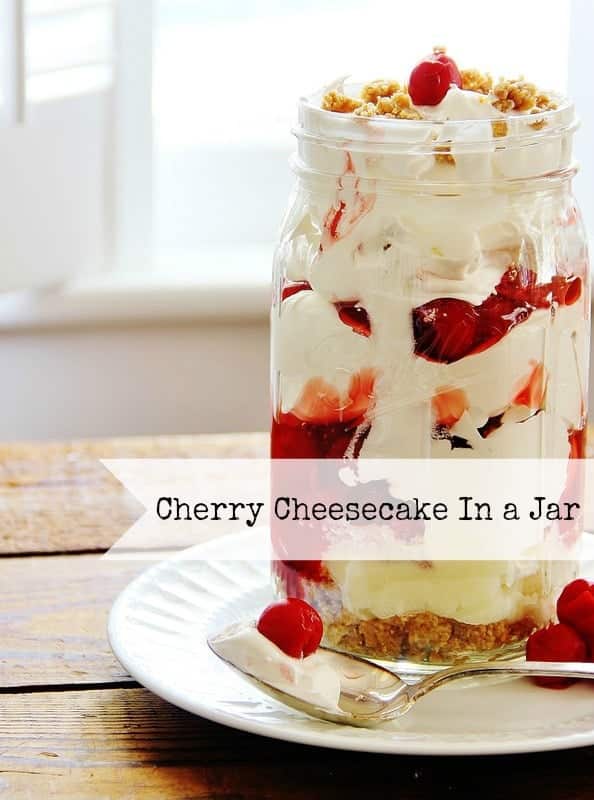 A mason jar filled with layers of cherry cheesecake, topped with cherries and a crumbly topping, perfect for festive gatherings. It's placed on a white plate next to a dollop of whipped cream and a cherry. A label reads "Cherry Cheesecake In a Jar," ideal for New Year's recipes.