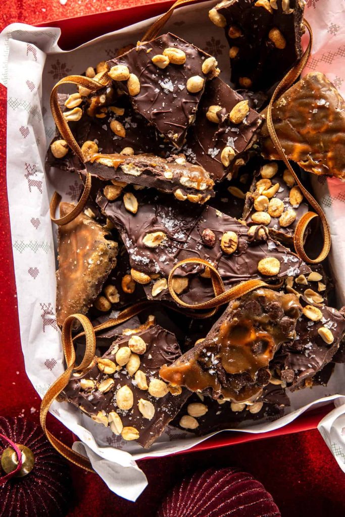 A festive box filled with chocolate bark topped with crushed peanuts and drizzled caramel, perfect for those seeking Christmas treat recipes. The chocolate pieces vary in size, artfully arranged among decorative ribbons. The box sits on a red surface with subtle holiday decorations.