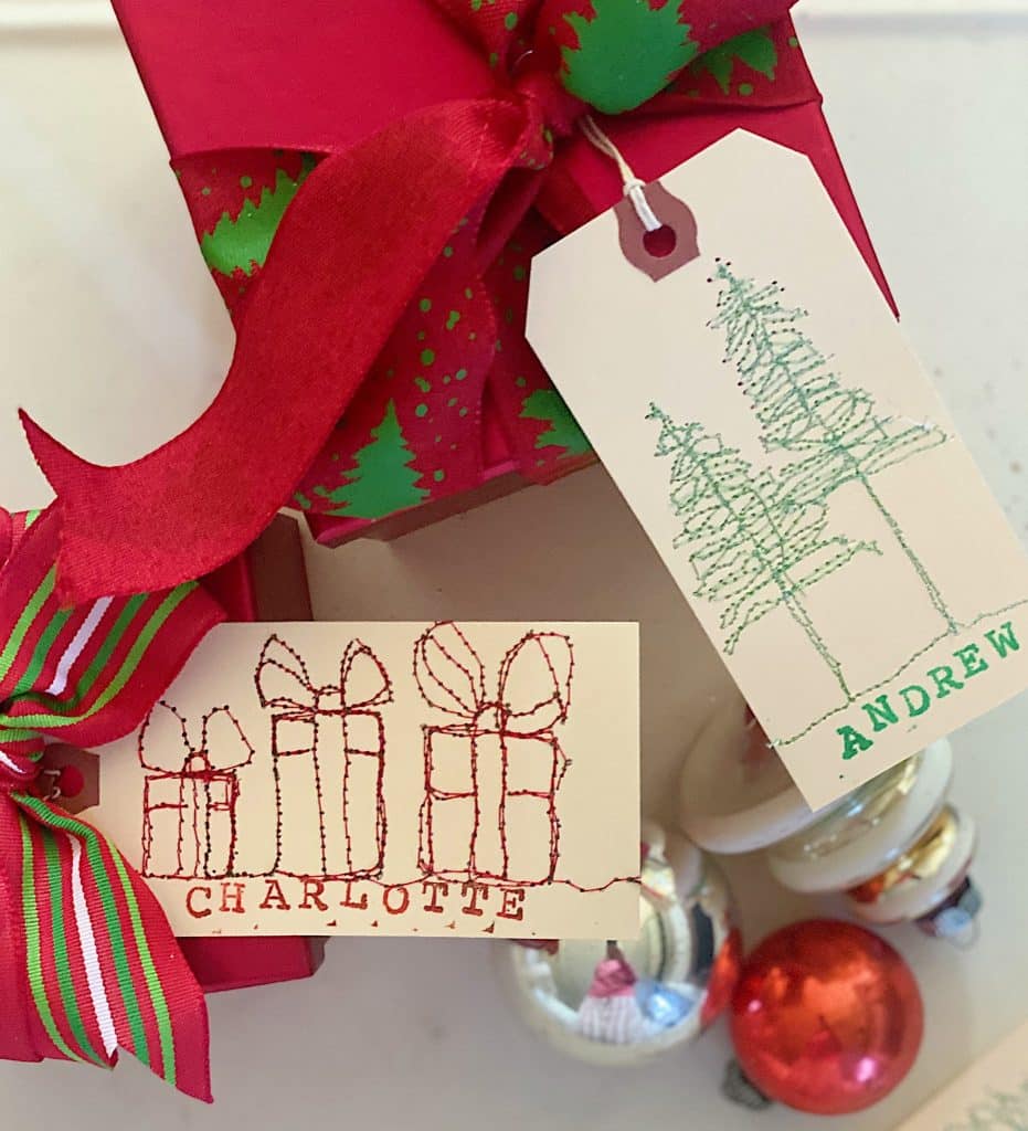 Two wrapped gifts feature festive red ribbons and green tree designs. Adorned with cute Christmas tags, one reads "Charlotte" with gift drawings, and the other says "Andrew" with tree illustrations. Decorative ornaments peek from the bottom.