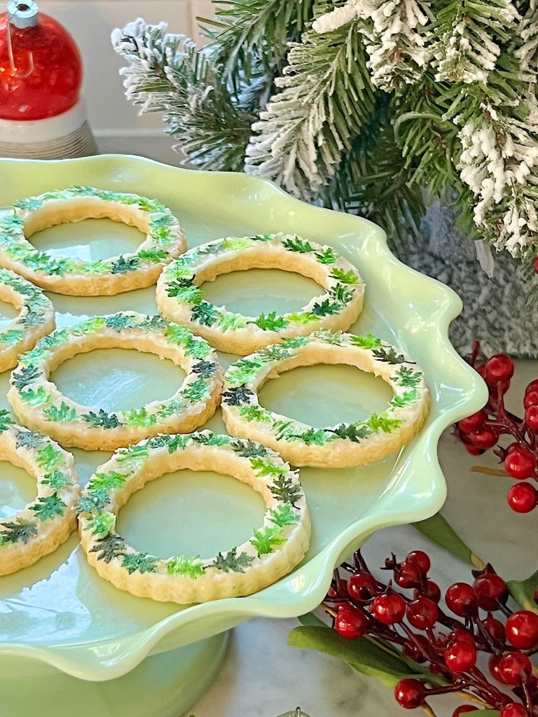 A plate of round decorative cookies with green and snow-like icing on the edges, resembling wreaths. Perfect for Christmas treat recipes, these cookies rest on a pale green stand. In the background, a frosted pine branch and red berries create a festive atmosphere.