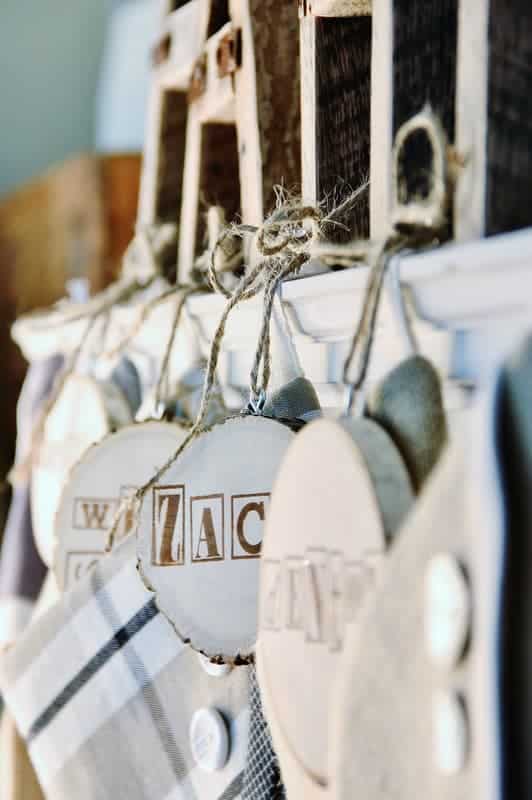 Close-up of handmade rustic decorations with wooden and fabric elements hanging on twine, resembling cute Christmas tags. Each circular piece features stenciled letters like "AC" and "NAP." A blurred background enhances the cozy, country-style aesthetic.