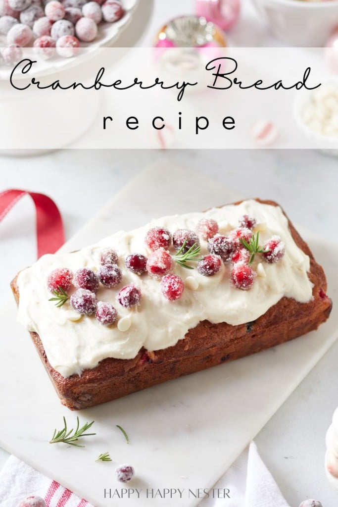 A loaf from our Cranberry Bread Recipe, topped with creamy frosting and sugared cranberries, sits elegantly on a white marble board. Sprigs of rosemary garnish the bread while baking ingredients and more sugared cranberries linger in the background, adding to the festive allure.