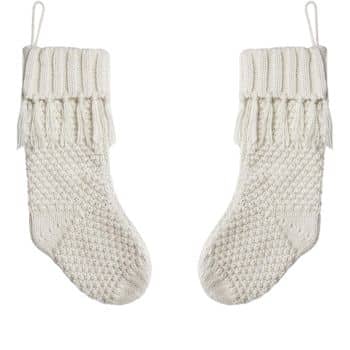 Two off-white knitted Christmas stockings hanging against a white background. Each stocking features a ribbed cuff and textured knit pattern with tassel details at the top.