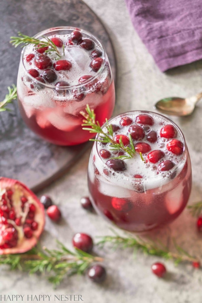 Two glasses of a festive drink, perfect for New Year's recipes, are garnished with cranberries, rosemary sprigs, and ice on a countertop. A pomegranate half and scattered cranberries add to the scene, while a purple napkin and spoon peek from the background.