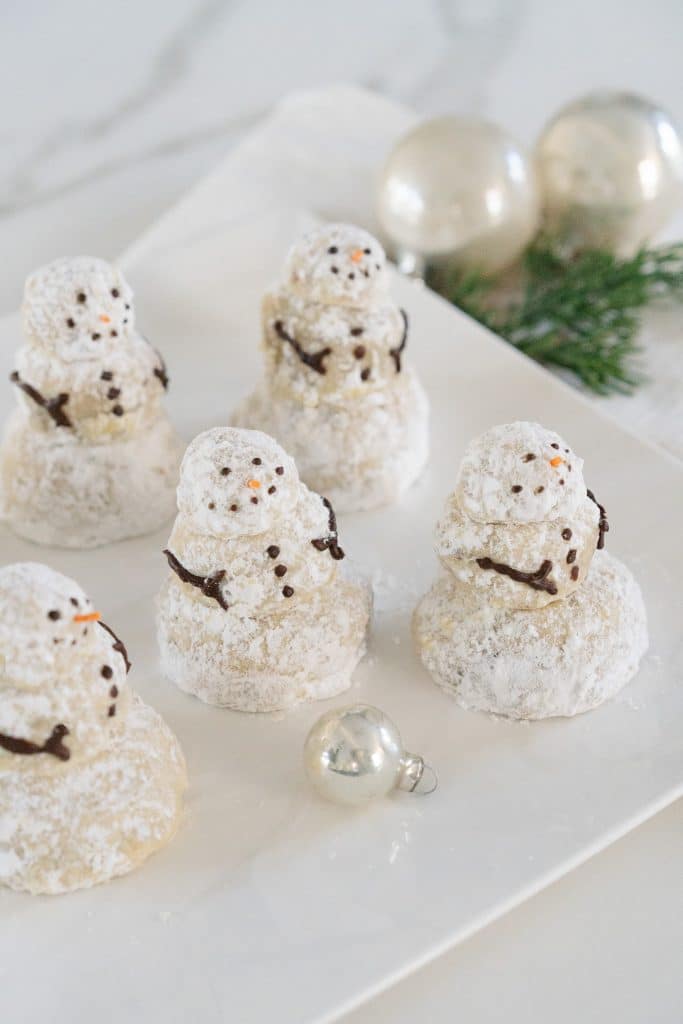 Five snowman-shaped powdered cookies arranged on a white plate evoke the festive spirit. Each cookie is decorated with chocolate eyes, buttons, and scarves, reminiscent of Christmas tree cookies. Silver ornament balls and green pine sprigs provide a cheerful backdrop.