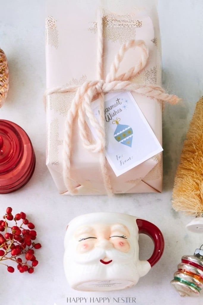 A festive scene with a wrapped gift featuring a star-patterned paper and a holiday card, a Santa-shaped mug, red berries, a red glass ornament, and a straw tree decoration on a light background.