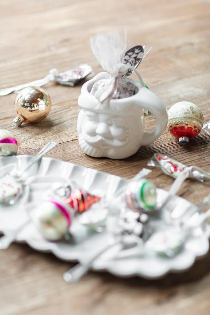 Festive scene with assorted Christmas ornaments and vintage-style lollipops on a wooden table. A Santa-shaped mug holds lollipops, surrounded by shiny baubles, snowglobe cookies, and a decorative tray.