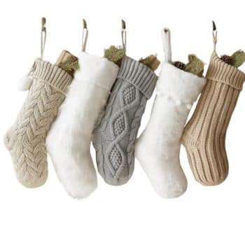 Five Christmas stockings hanging in a row: two are knit in beige, one white and fluffy, one knit in gray with diamond patterns, and another white and fluffy. Each is filled with pine cones and greenery.