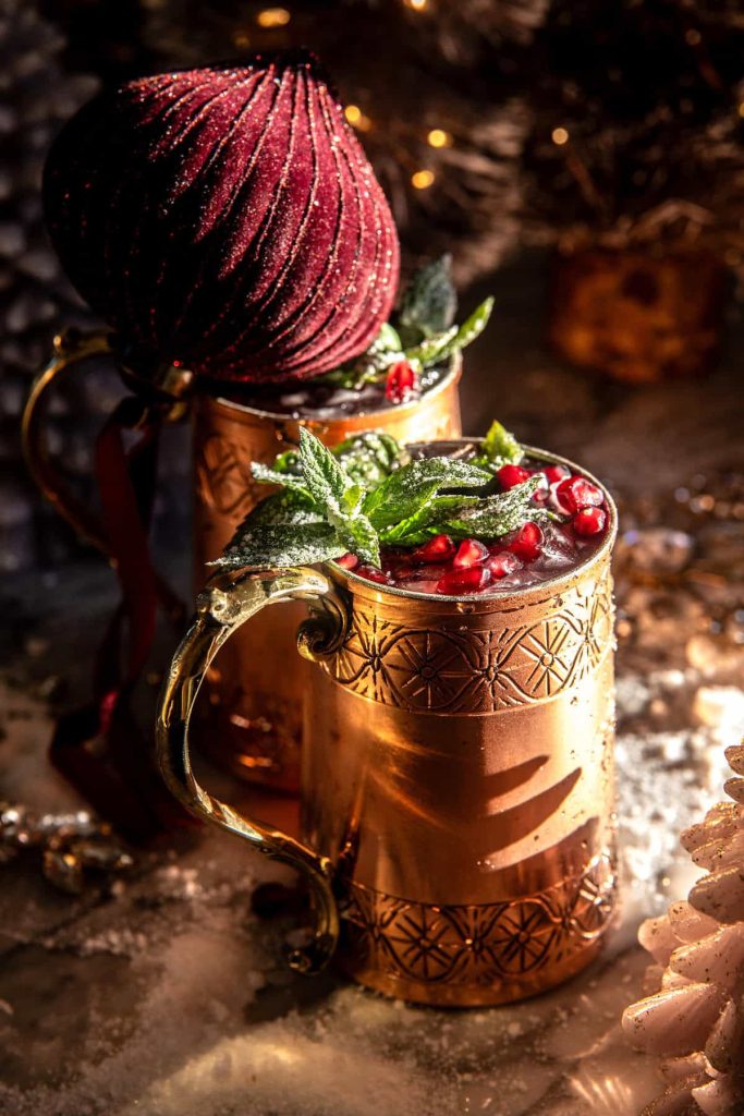 Festive copper mugs filled with cranberry drinks, garnished with mint leaves and pomegranate seeds, sit on a snowy surface. A large burgundy Christmas ornament rests on the mug handle, creating a cozy holiday scene with warm, glowing lights—perfect for inspiring new year's recipes.