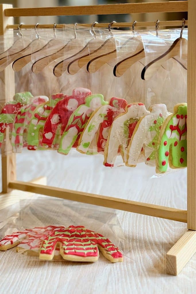 A wooden rack displays creatively decorated Christmas tree cookies, alongside other sweater-shaped treats, each packaged in clear plastic and hanging on mini hangers. The cookies feature festive designs in red, green, and white with various patterns.