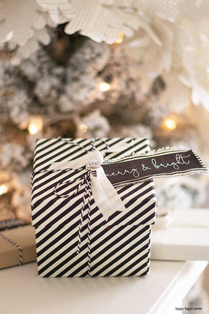 A gift wrapped in black and white striped paper with a cute Christmas tag reading "merry & bright" hangs from a white ribbon. It sits gracefully in front of a blurred Christmas tree adorned with twinkling lights.