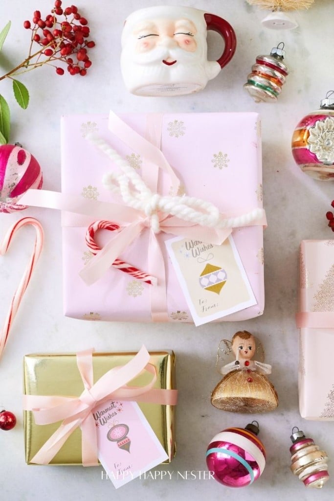 A festive holiday scene unfolds with wrapped gifts, a Santa-themed mug, vintage ornaments, berries, and a candy cane on a white surface. One gift is adorned in pink paper with a white bow and another in gold with a pink ribbon, accompanied by free Christmas tags printable for adding personal touches.