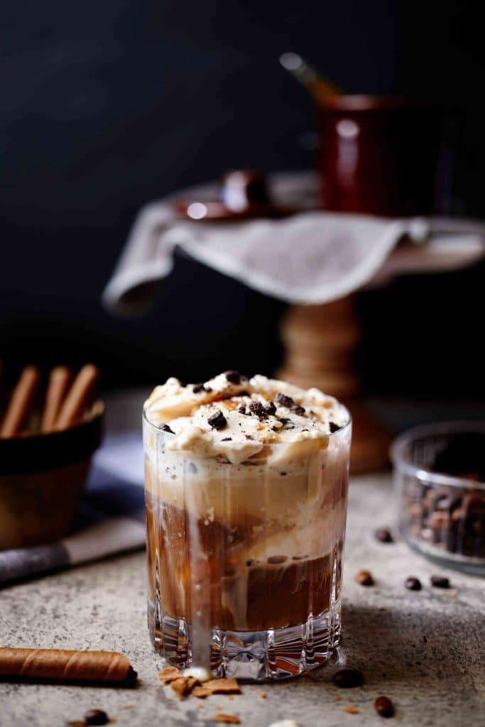 A glass of creamy iced coffee, perfect for New Year's recipes, is topped with whipped cream and chocolate shavings on a rustic surface. Nearby are coffee beans, cinnamon sticks, and a wooden stand with a draped cloth and small pot in the background.