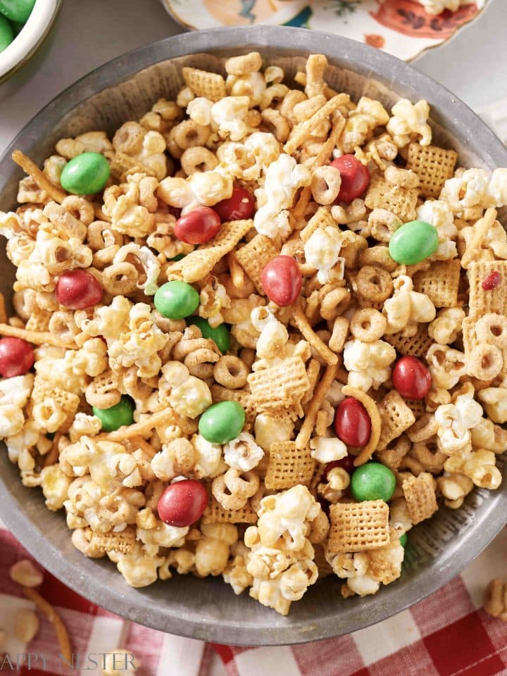 A bowl filled with a delightful Christmas treat mix featuring popcorn, red and green candy-coated chocolates, cereal pieces, pretzel sticks, and chex cereal. The festive blend offers a colorful array of textures perfect for the holiday season.