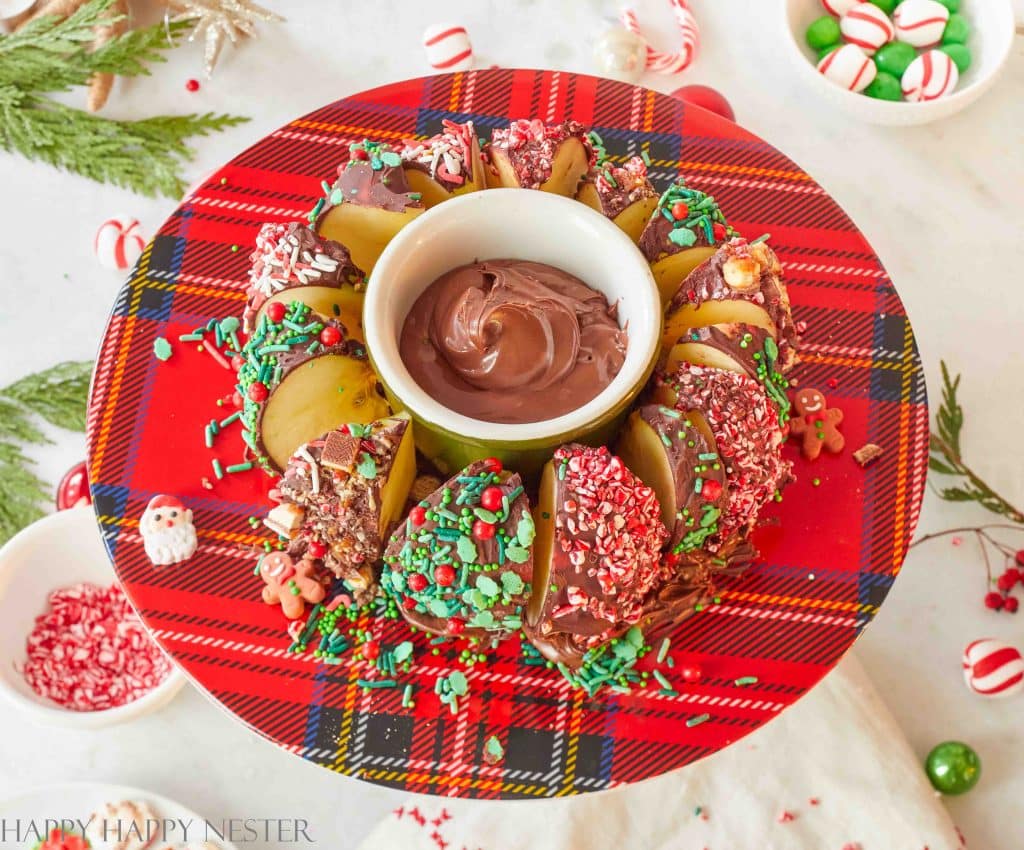 Festive apple slices covered in chocolate and colorful sprinkles are arranged in a circle on a red plaid plate. A small bowl of chocolate dip is in the center, making it a perfect addition to your Christmas treat recipes. Holiday-themed decorations surround the plate.