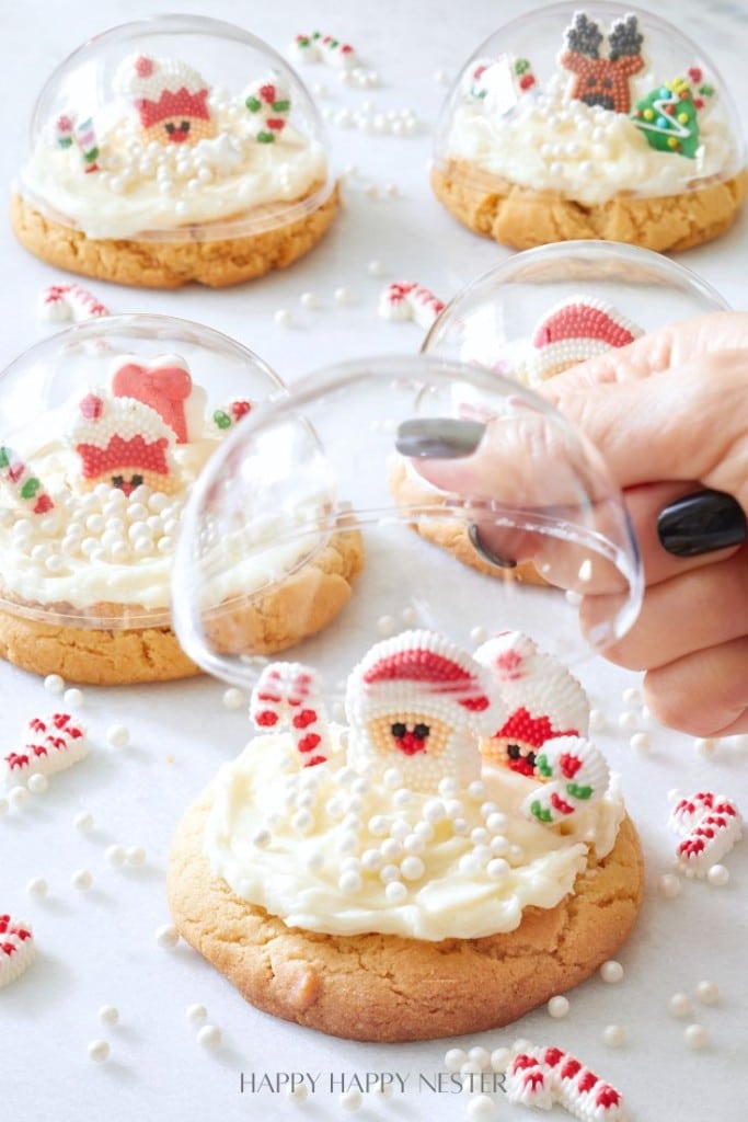 A person places a clear dome over a beautifully decorated cookie, transforming it into an enchanting Snow Glove Cookie. The cookie is topped with white frosting, a small Santa figure, and edible pearls. Similar cookies are in the background amidst festive decorations and scattered sprinkles.